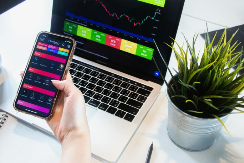 A person using a smartphone app to trade stocks with a laptop displaying market data.