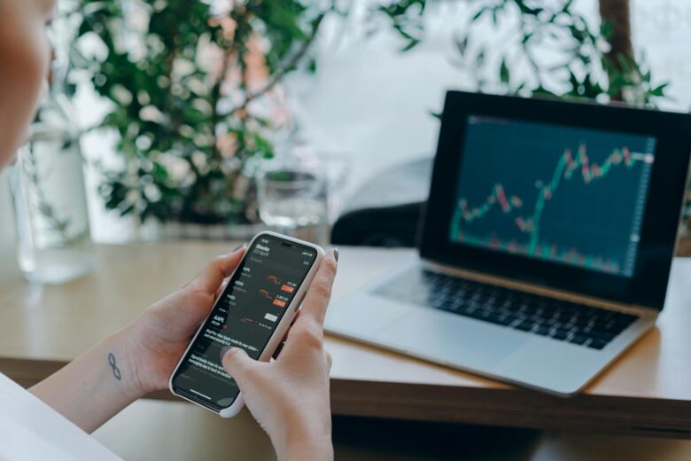 Businesswoman reviewing financial data on smartphone alongside laptop displaying graphs in modern workspace.
