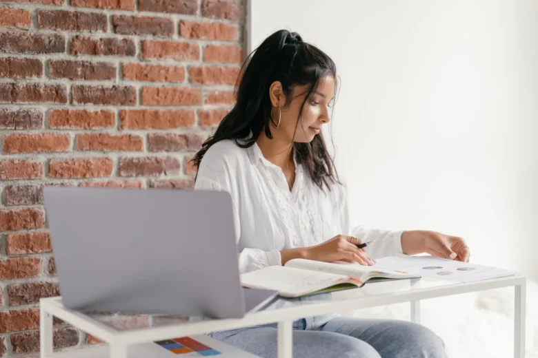Empreendedora trabalha em uma mesa doméstica com notebook e caderno de anotações, representando um negócio iniciado com pouco investimento